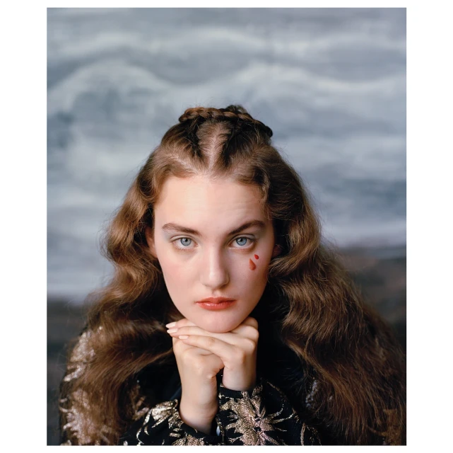 Portrait of a young woman with auburn hair and a braided crown, resting her chin on her clasped hands. A single red tear marks her cheek.
