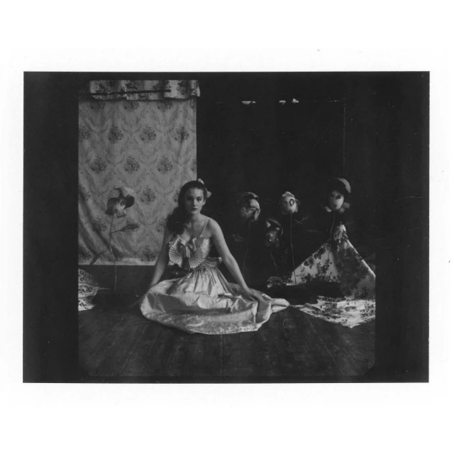 Monochrome photograph of a seated woman in a light dress, flanked by mannequin heads and decorative floral wallpaper in the background.
