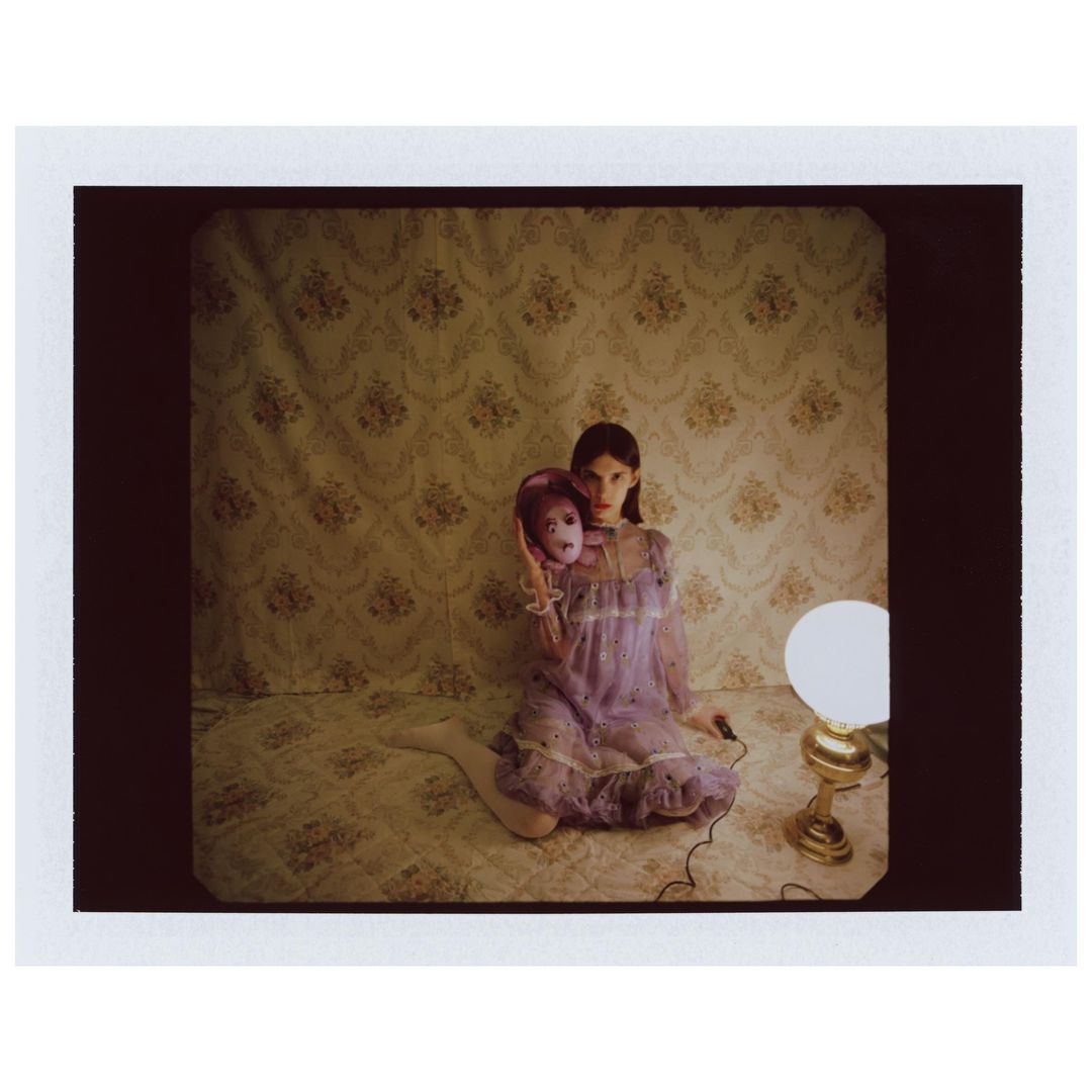 A woman in a lilac dress sits on a bed, holding a doll-like mask, in a room with floral wallpaper and a vintage lamp.