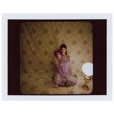 Woman in sheer purple dress holding purple mask seated next to a lit lamp on floral wallpaper background