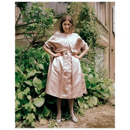 Woman in shiny pink dress with hands on hips standing outdoors before lush green foliage and a stone building.