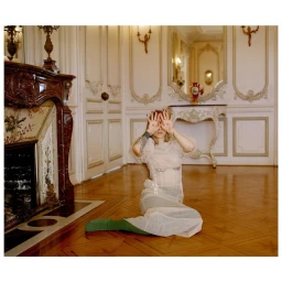 Woman in sheer white dress sitting on a wooden parquet floor in a classical interior with a marble fireplace and ornate wall panels.