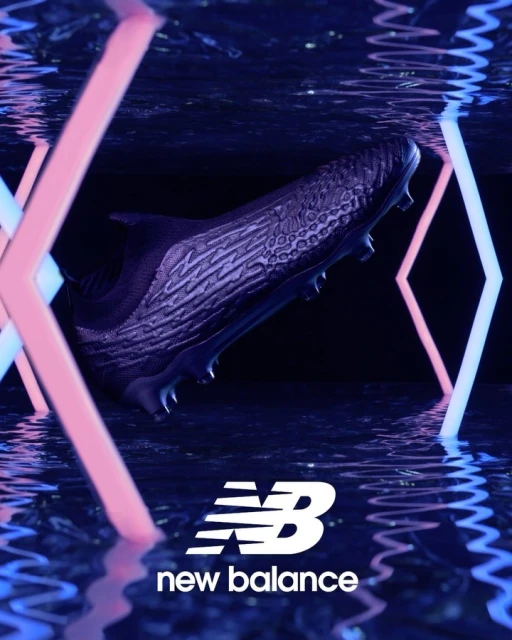 Close-up of a black New Balance soccer shoe against a dark backdrop with blue and pink neon light accents, highlighting the brand logo.