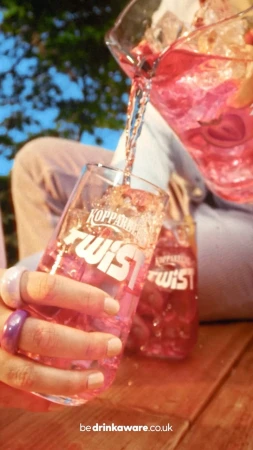 Close-up of a hand pouring a strawberry-infused drink from a pitcher into a glass with a Kopparberg Twist logo; a second glass sits nearby.