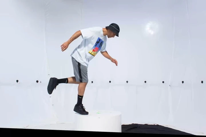 Man in casualwear jumps off platform in studio setting.  He wears a tee shirt, shorts, bucket hat, and sneakers, poised mid-air.