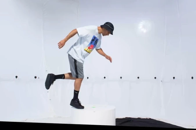 Man in casualwear jumps off platform in studio setting.  He wears a tee shirt, shorts, bucket hat, and sneakers, poised mid-air.