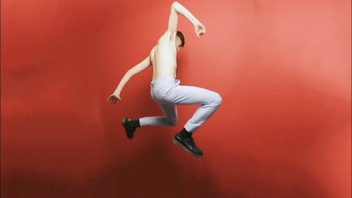 An adult male is leaping in the air in an indoor setting, wearing light blue pants and black shoes, against a red wall background.