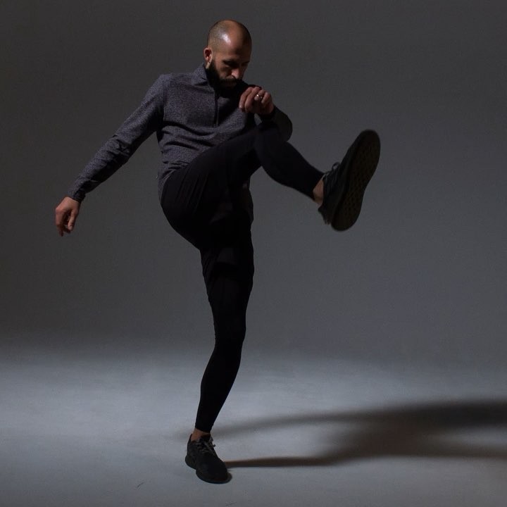 Athletic bald man strikes a dynamic pose in a gray studio, wearing black pants and a gray quarter-zip top, with a shadow on the floor.