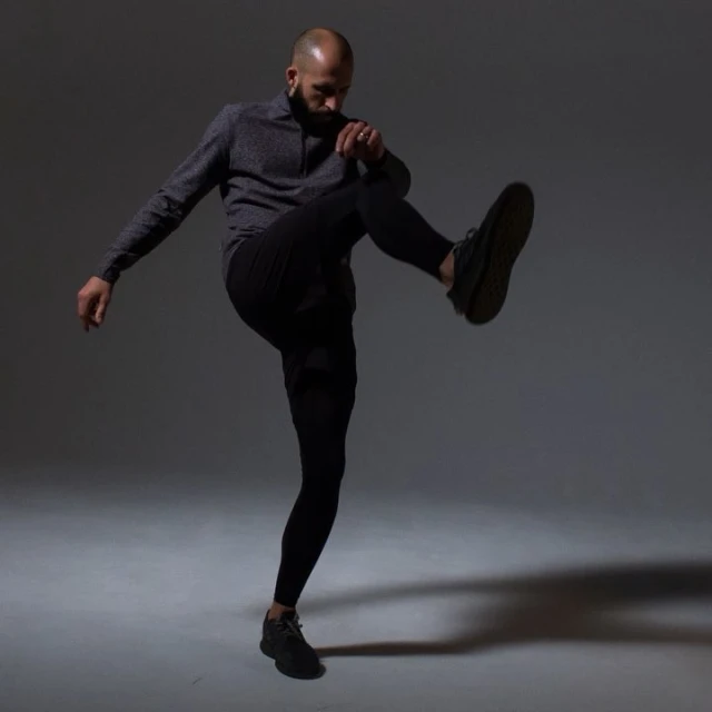 Athletic bald man strikes a dynamic pose in a gray studio, wearing black pants and a gray quarter-zip top, with a shadow on the floor.