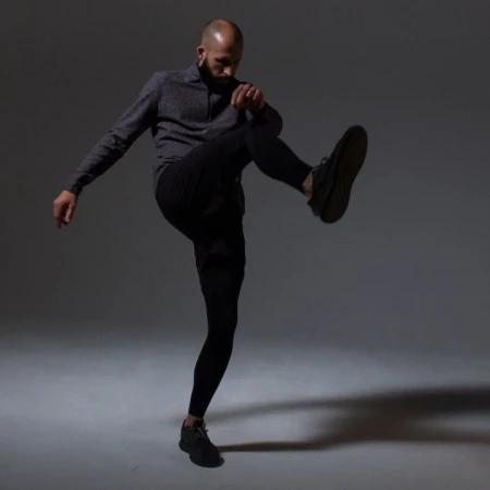 Athletic bald man strikes a dynamic pose in a gray studio, wearing black pants and a gray quarter-zip top, with a shadow on the floor.