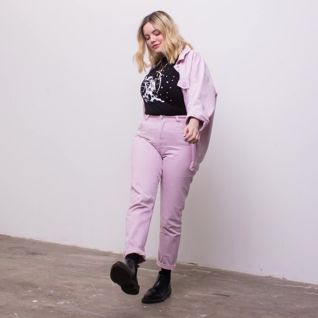 Blonde woman in a black graphic tee, pink pants, and jacket, walking indoors with a white wall backdrop and concrete floor.