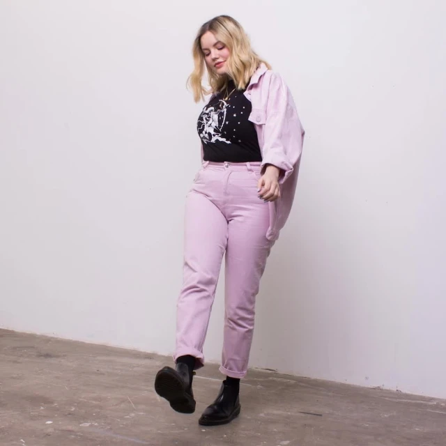Blonde woman in a black graphic tee, pink pants, and jacket, walking indoors with a white wall backdrop and concrete floor.
