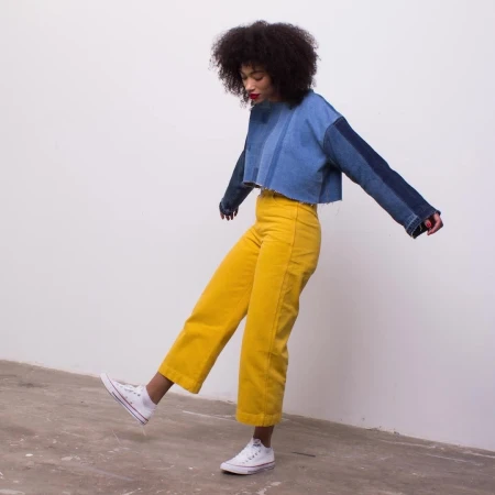 A young adult female with afro-style hair strikes a pose in a studio, wearing a patchwork denim top and mustard-yellow wide-leg pants.