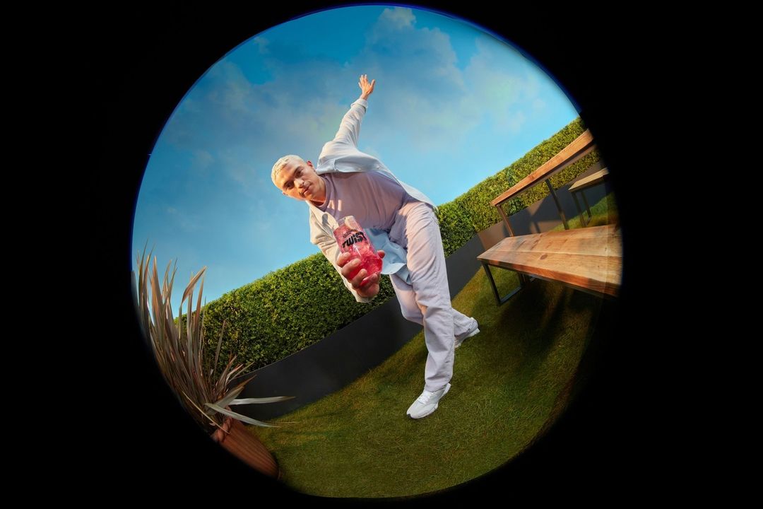 Person in white suit poses with pink bottle on lawn under sky, captured with fisheye lens and outdoor setting.