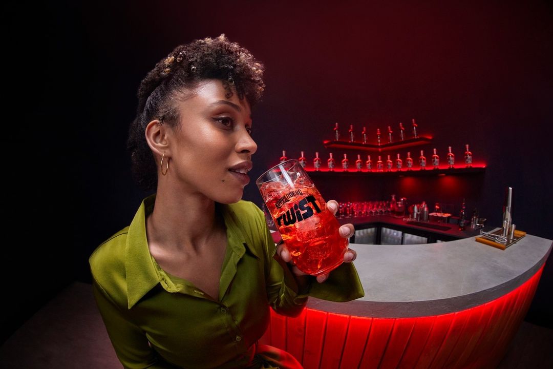 A person with curly hair, in a green top, drinks a red cocktail from a branded glass at a red-lit bar, offering a vibrant and inviting scene.