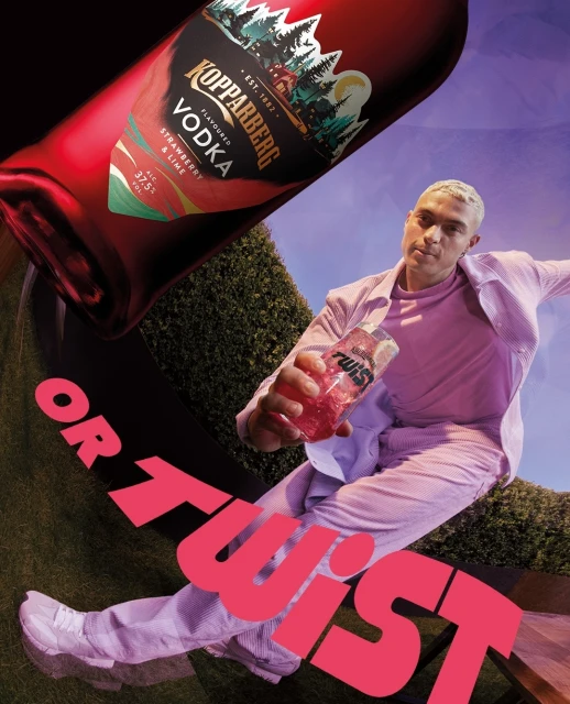 Man in a pink suit holds a Kopparberg Twist drink, with a large Kopparberg Vodka bottle behind him on a green and blue background.