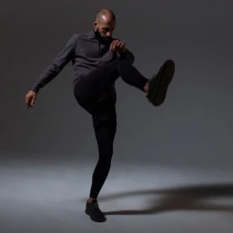 A bald man with a beard kicking his leg up while wearing a dark grey shirt, black pants, and dark sneakers in a studio.