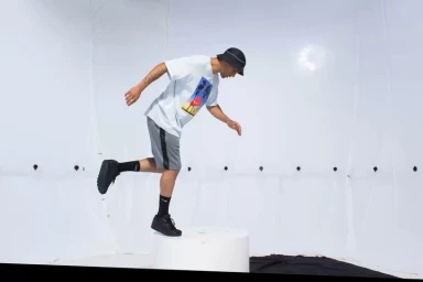 Man wearing a white t-shirt, grey shorts, and bucket hat stepping onto a white platform in a seamless white studio.