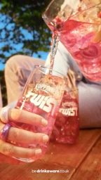 Pink beverage being poured from a pitcher into a tall branded glass filled with ice by a person wearing rings
