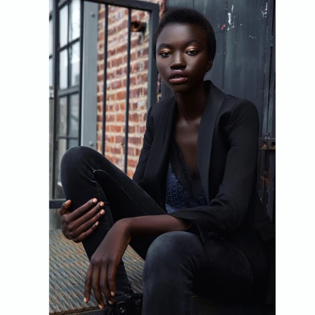 Portrait of a dark-skinned woman in black blazer and dark jeans sitting on steps, with a brick wall and dark door in the background.