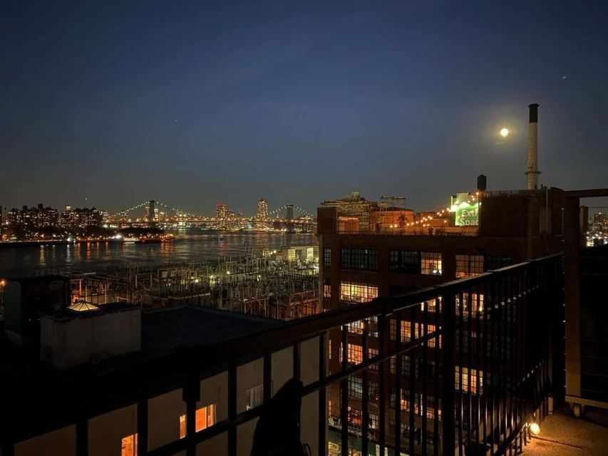 Cityscape at night with a view of bridges and buildings across a river, lit by lights, under a dark sky with a full moon.