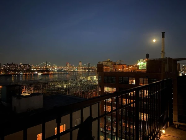 Cityscape at night with a view of bridges and buildings across a river, lit by lights, under a dark sky with a full moon.