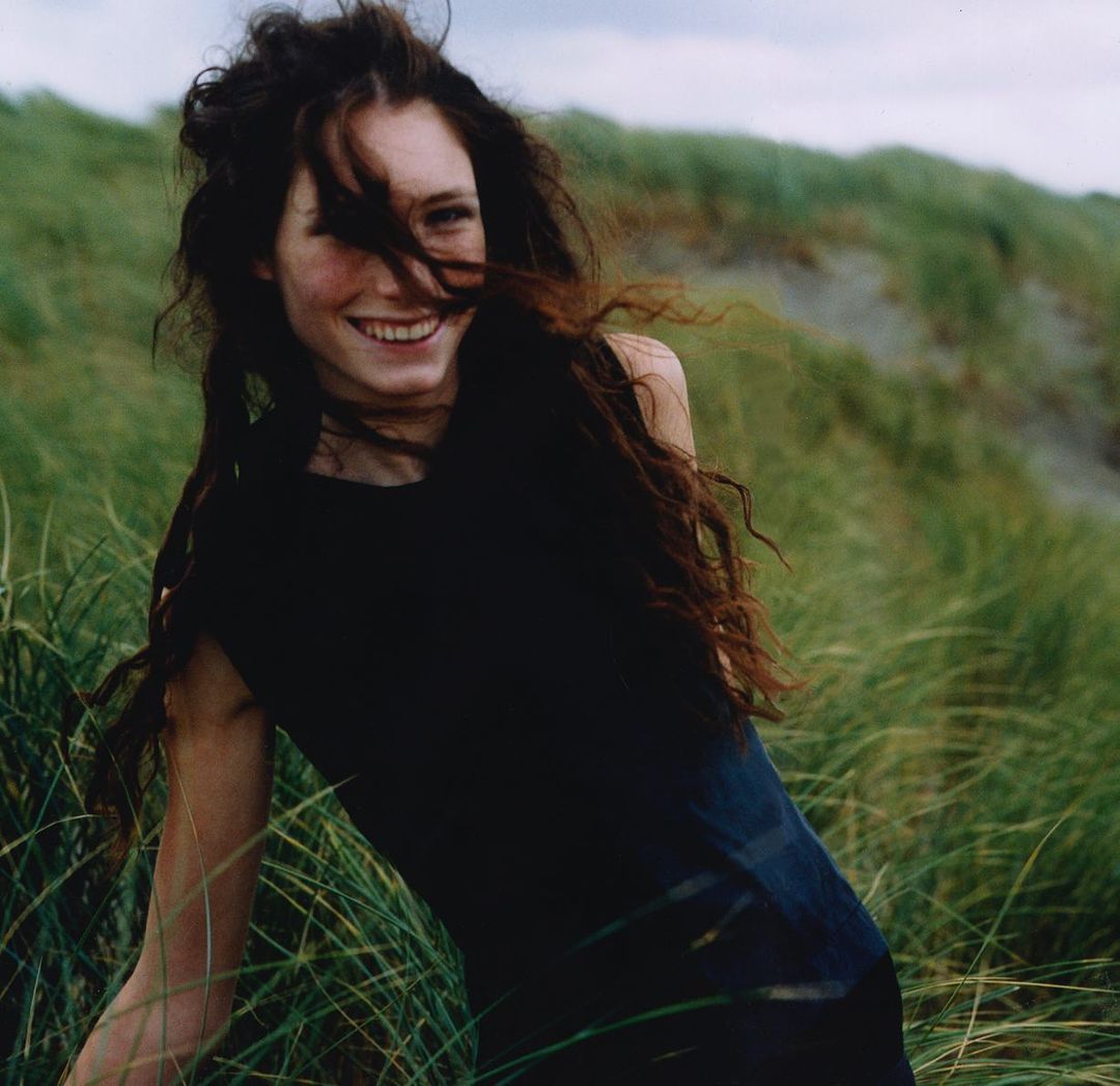 Smiling woman with long hair, wearing black top, posing in tall green grass in front of a grassy hill on a sunny day outdoors.