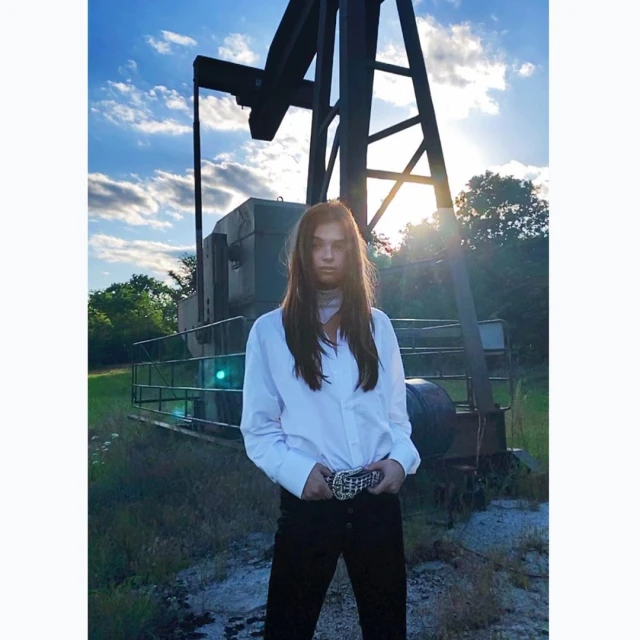 Adult female poses outdoors in white blouse and black pants near an oil well against blue sky and clouds during daytime hours.