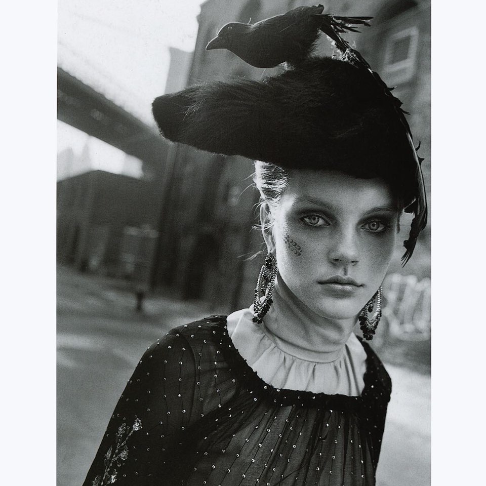Monochrome portrait of a woman with a black hat adorned with a bird, wearing beaded earrings and a dress with a city background.