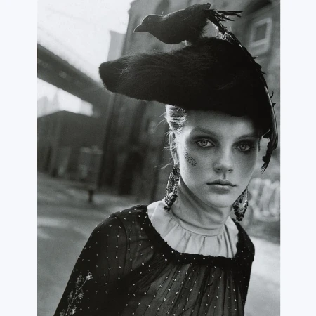 Monochrome portrait of a woman wearing a black taxidermy bird hat and beaded earrings against an urban street background.