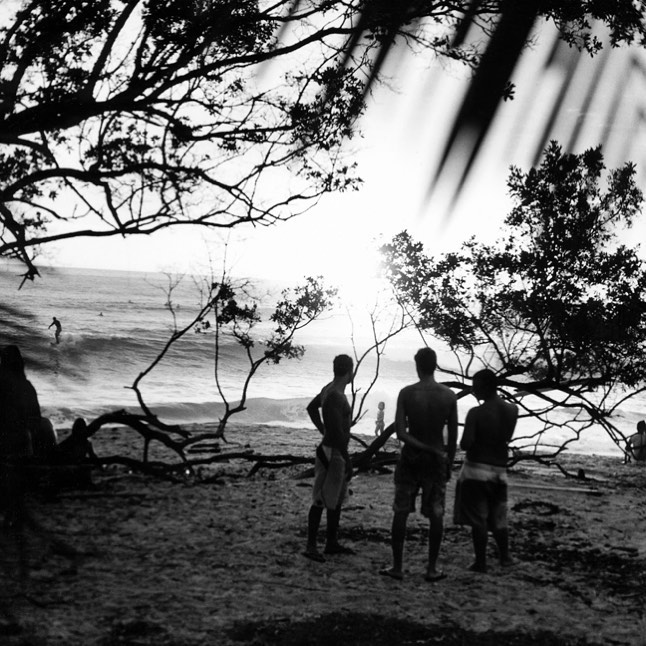Black and white beach scene featuring three men standing on the sand, ocean waves, silhouetted trees, and a surfer on the water.