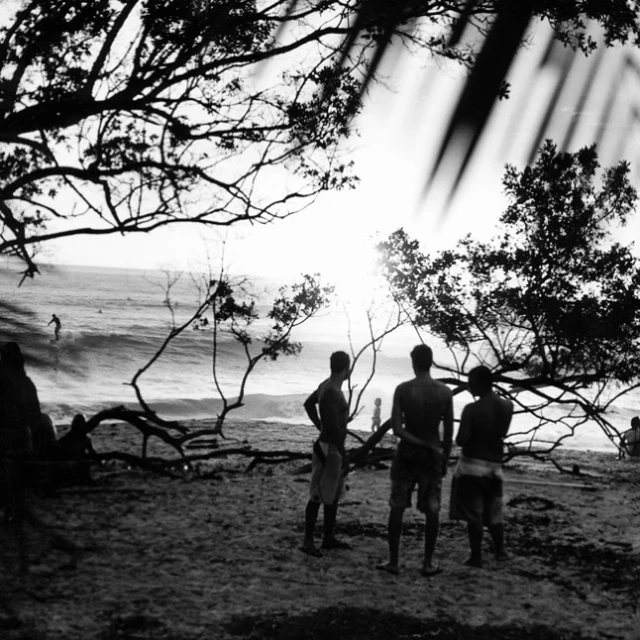 Black and white beach scene featuring three men standing on the sand, ocean waves, silhouetted trees, and a surfer on the water.