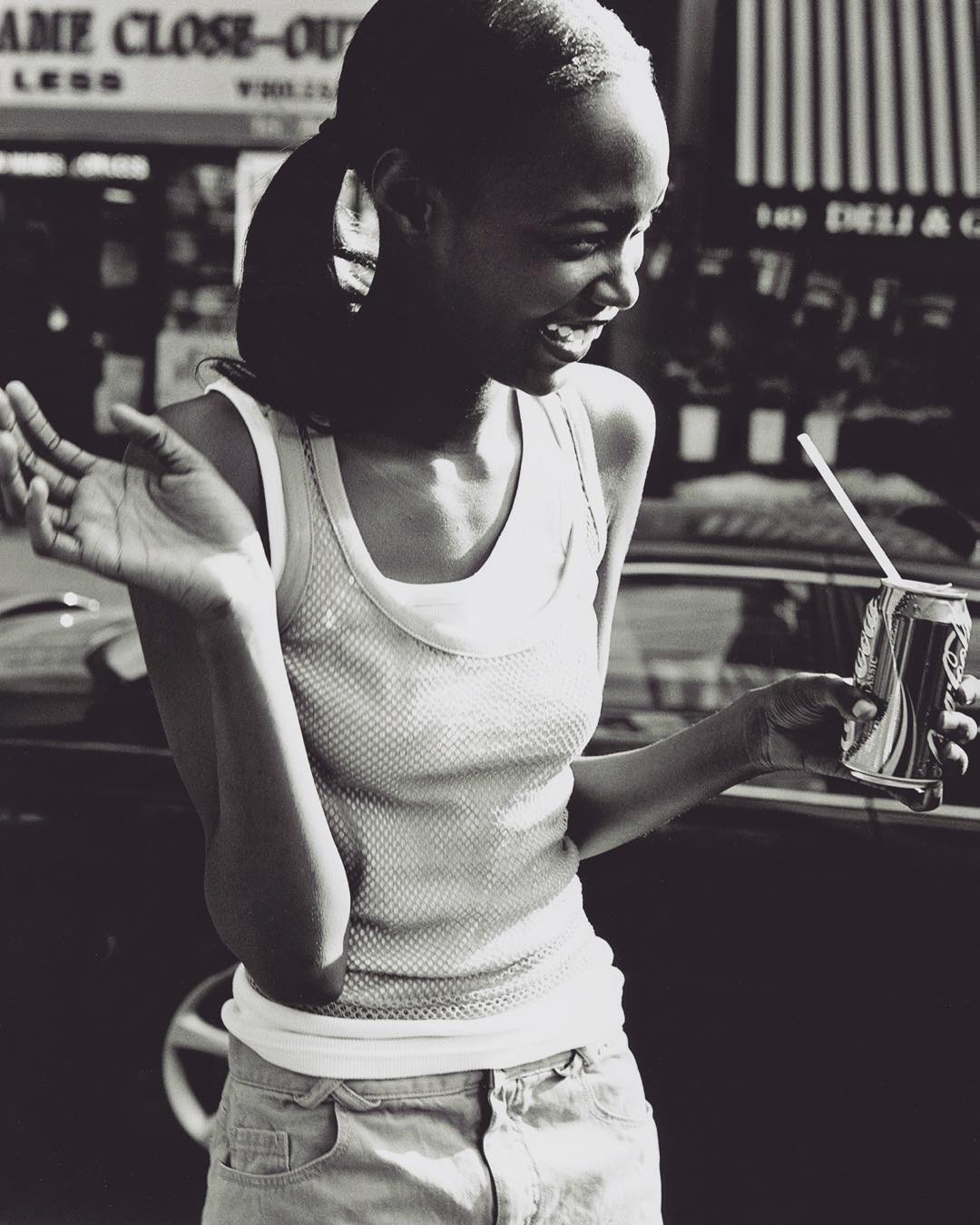 A happy, young woman smiles while holding a drink can with a straw. She wears a tank top and denim shorts on an outdoor city street.