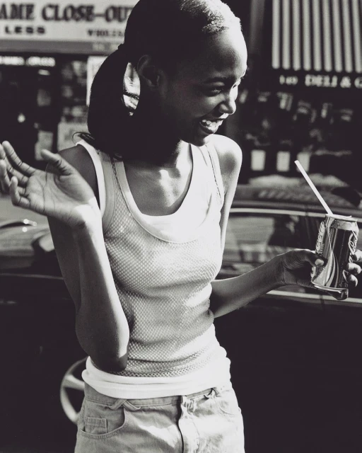 A happy, young woman smiles while holding a drink can with a straw. She wears a tank top and denim shorts on an outdoor city street.
