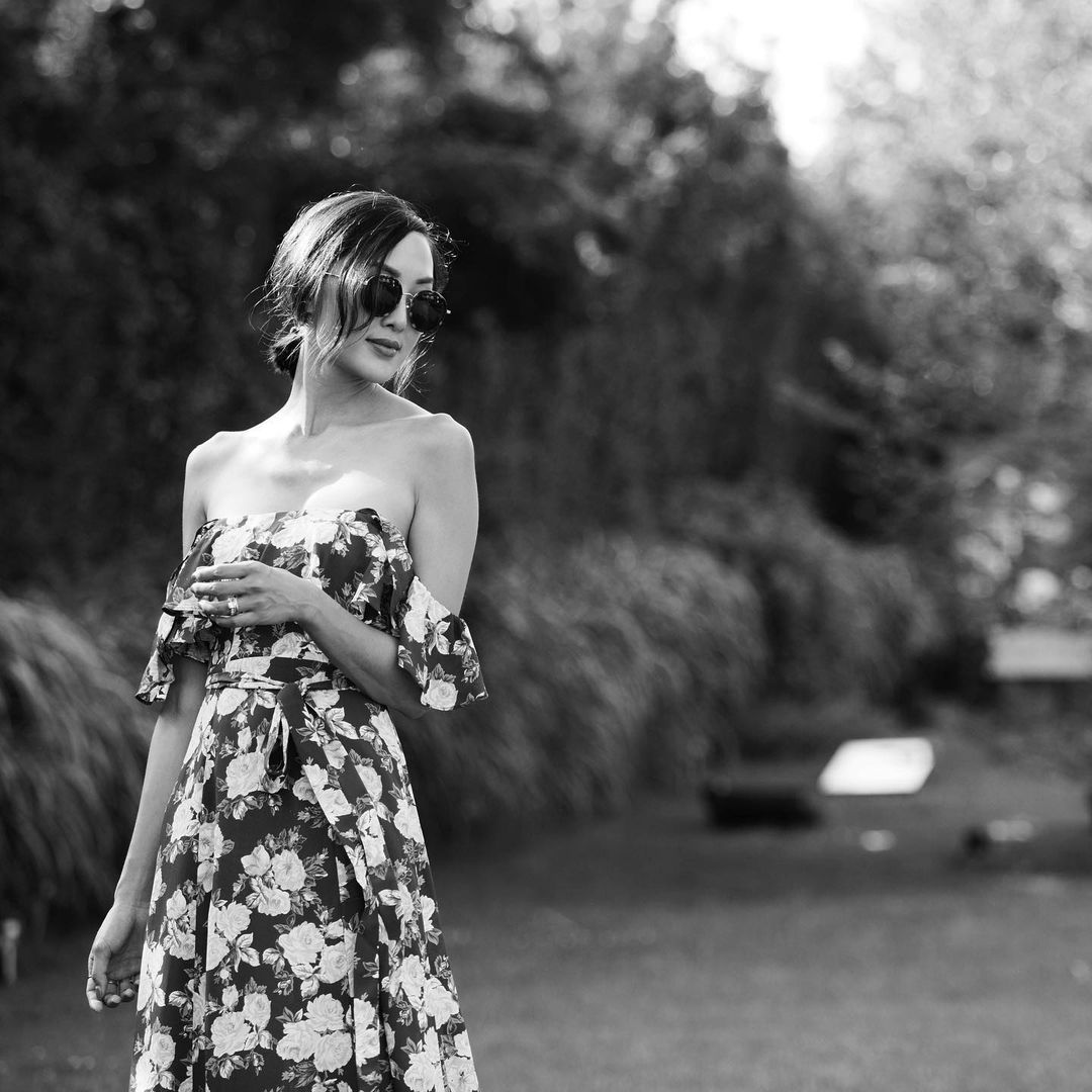 Black and white portrait of an Asian woman in a floral dress and sunglasses, posing outdoors against a natural green backdrop.