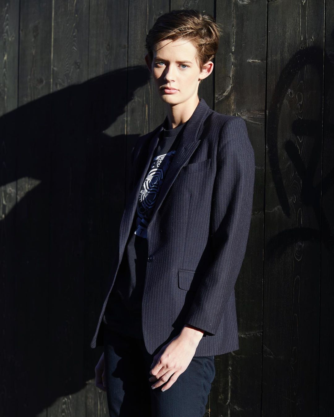Adult female in a blazer stands in front of a dark wall with shadows, creating a strong contrast and minimalist fashion-forward aesthetic.