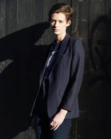Adult female in a blazer stands in front of a dark wall with shadows, creating a strong contrast and minimalist fashion-forward aesthetic.