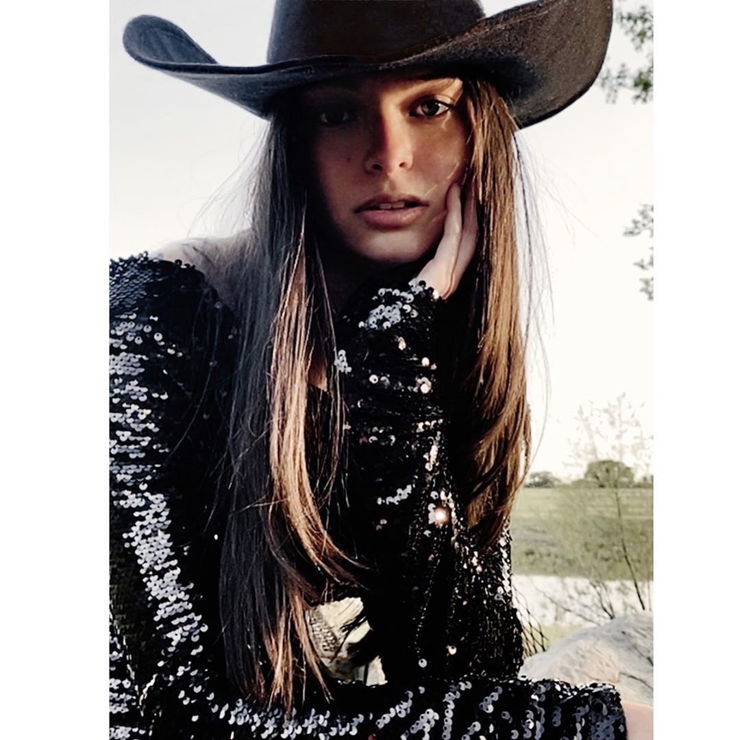 Woman in sequin dress and cowboy hat poses outdoors. Medium shot of female model in natural lighting against a bright background.