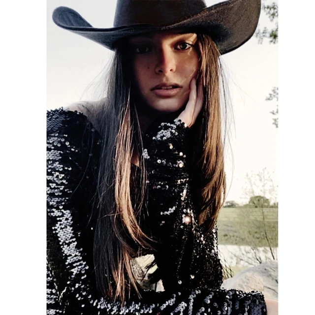 Woman in sequin dress and cowboy hat poses outdoors. Medium shot of female model in natural lighting against a bright background.