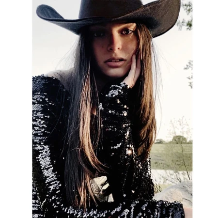 Woman in sequin dress and cowboy hat poses outdoors. Medium shot of female model in natural lighting against a bright background.