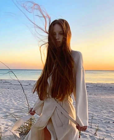 Woman with long brown hair blowing in the wind wearing a cream robe on a sandy beach during sunset.