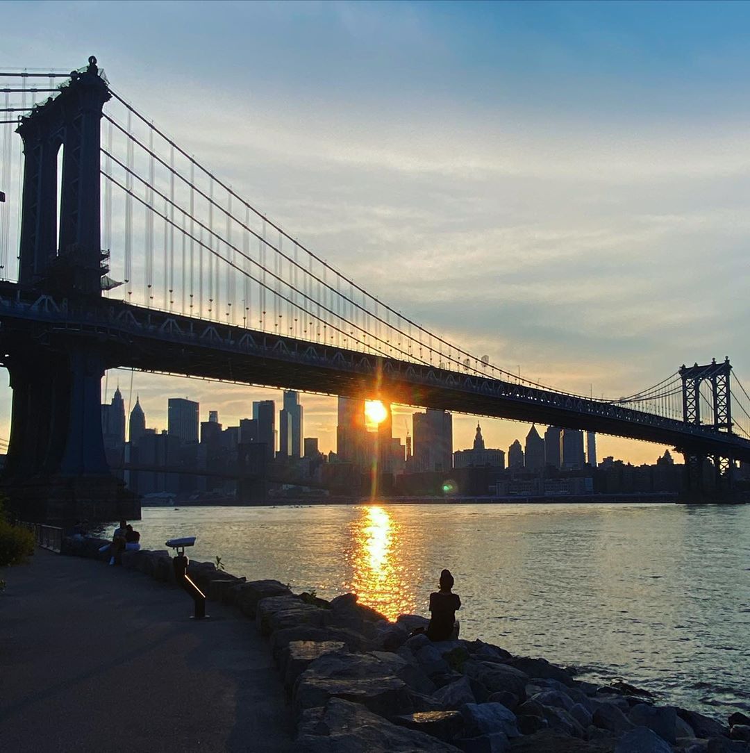 Manhattan Bridge view with the sun setting behind the city skyline, silhouetting the skyscrapers and bridge, with a calm river.