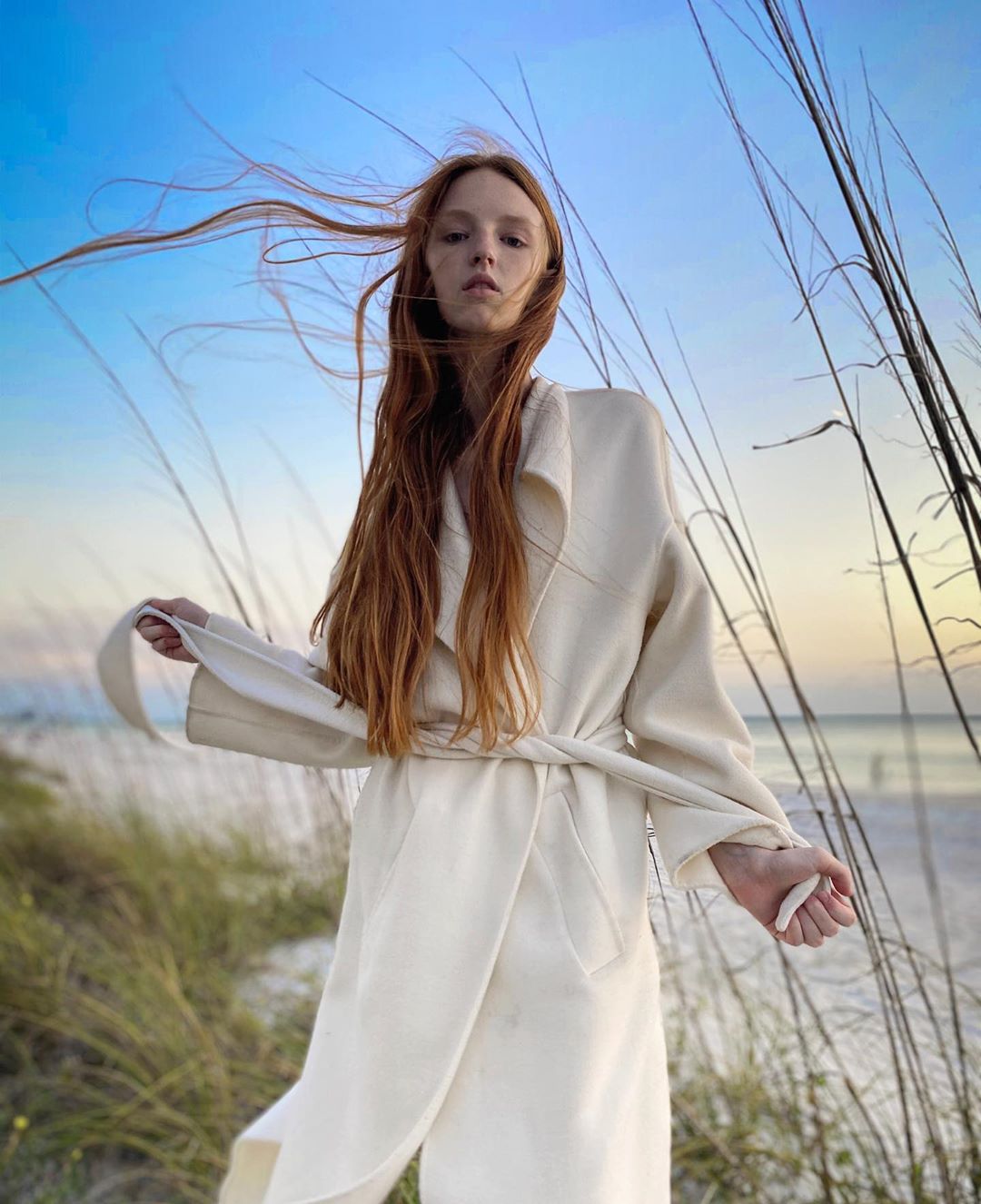 Woman with long red hair poses in a light beige coat on a beach with tall grass against a blue sky at dusk, fashion portrait.