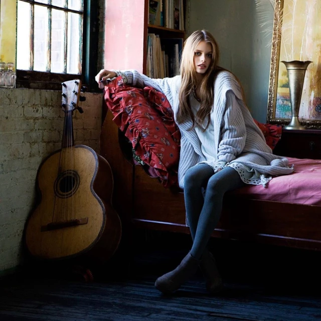 Young woman in gray sweater and tights sitting on bed, with guitar, window light, books, and vintage setting, indoor fashion.