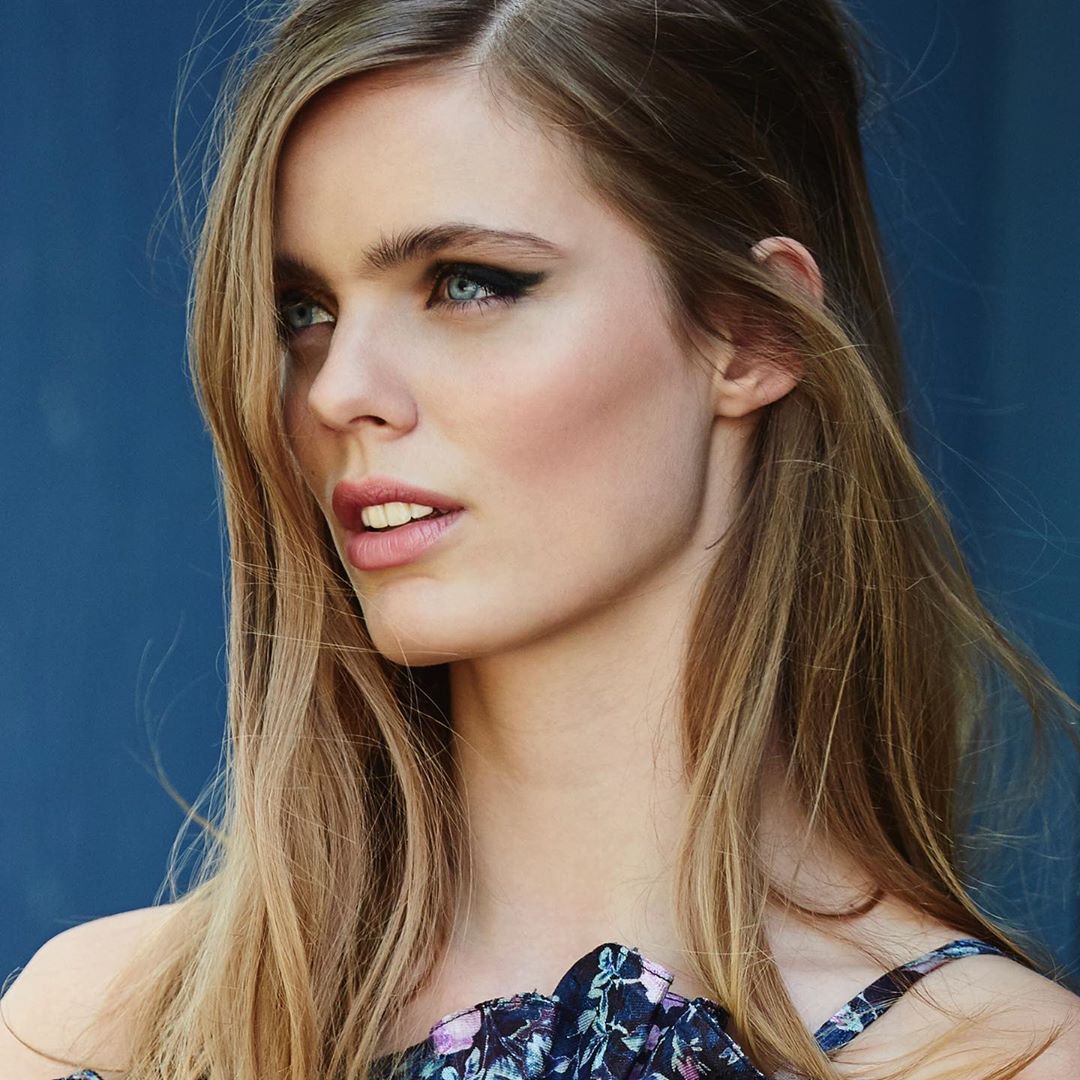 Close-up portrait of a young adult woman with blonde hair and blue eyes, wearing a floral top against a dark blue background.