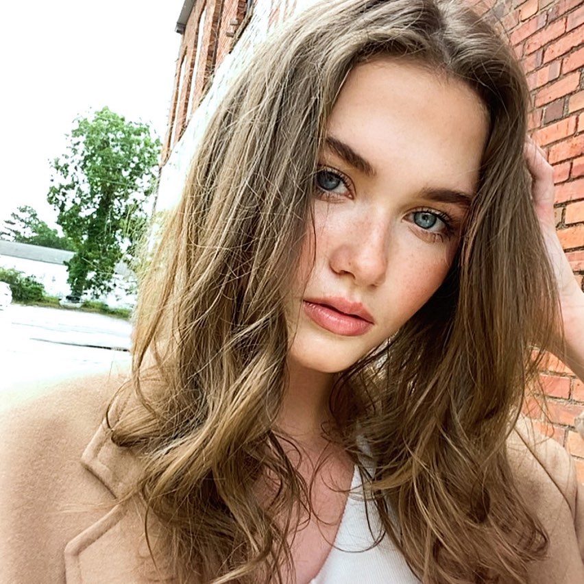 Close-up portrait of a young adult woman with wavy brown hair and blue eyes, wearing a beige coat, posing outdoors in daylight.