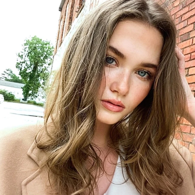 Close-up portrait of a young adult woman with wavy brown hair and blue eyes, wearing a beige coat, posing outdoors in daylight.