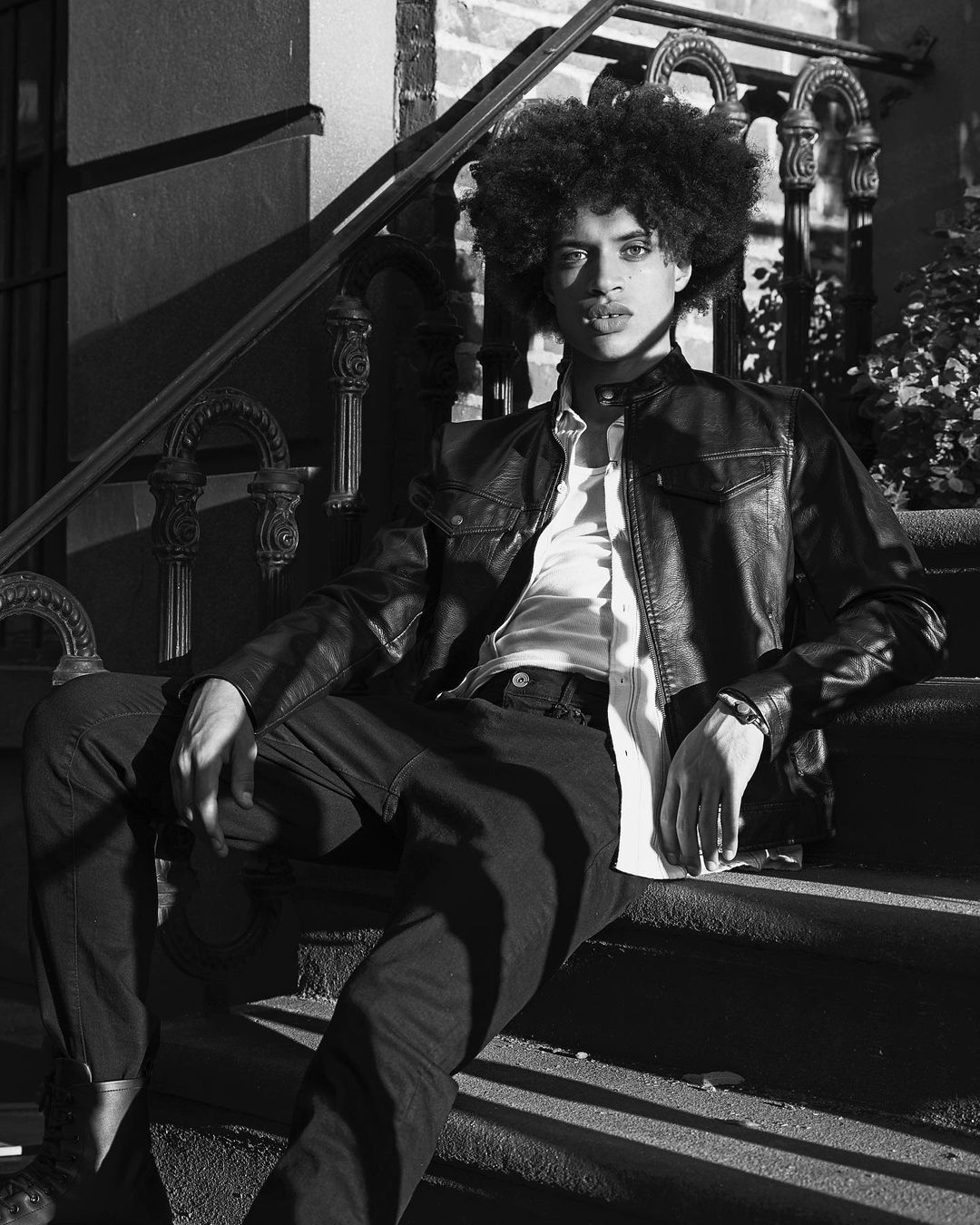 Black and white portrait of a young adult male with afro sitting on outdoor steps wearing a leather jacket, button-down shirt, and pants.