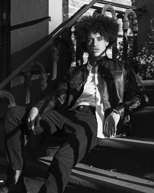 Black and white portrait of a young adult male with afro sitting on outdoor steps wearing a leather jacket, button-down shirt, and pants.