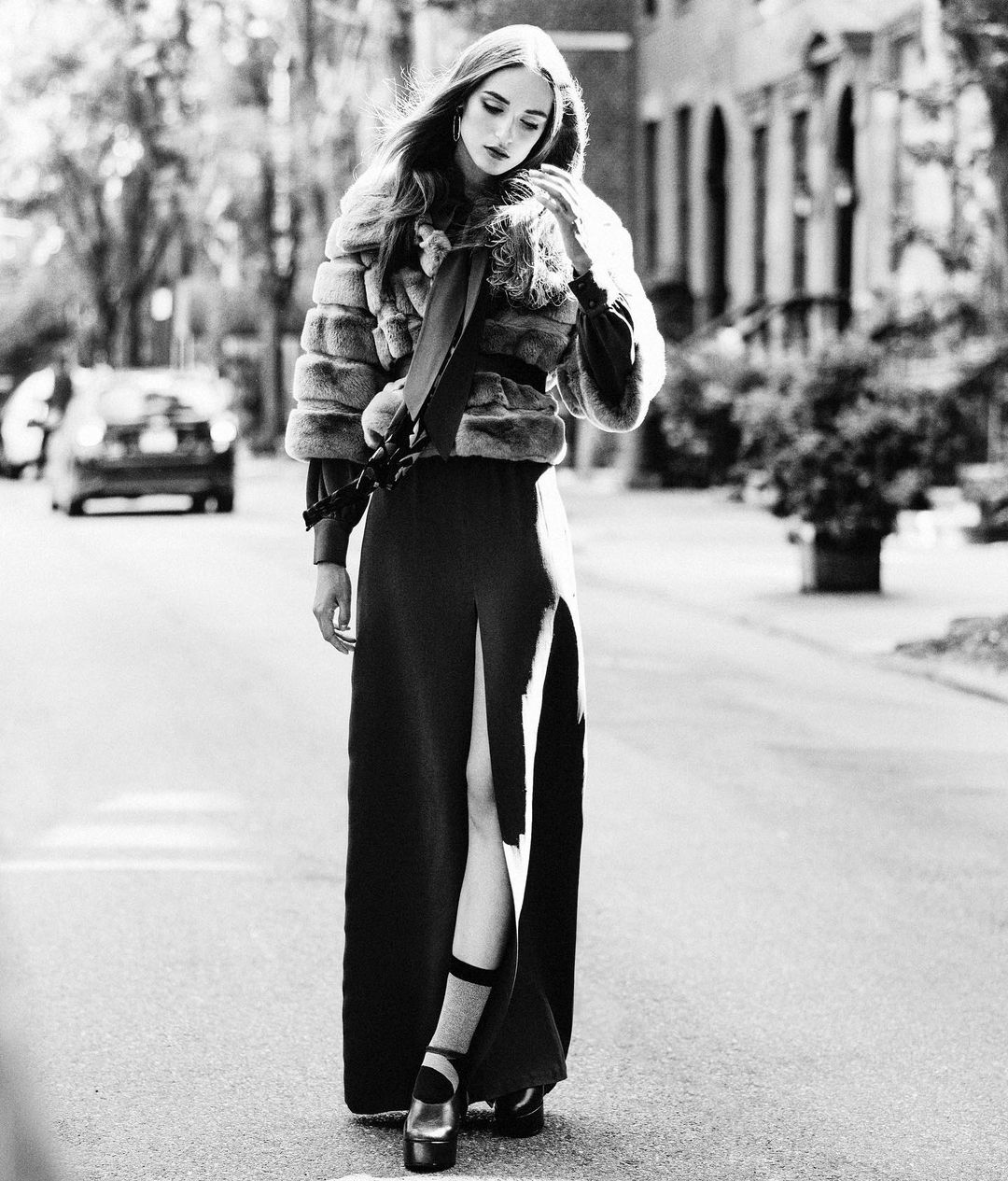 A young woman poses on a street in a black-and-white fashion shot, wearing a fur coat and a long black dress with a thigh-high slit.